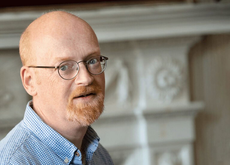 Portrait of a man with glasses and a red beard, wearing a blue checkered shirt, in an indoor setting with a decorative fireplace in the background.